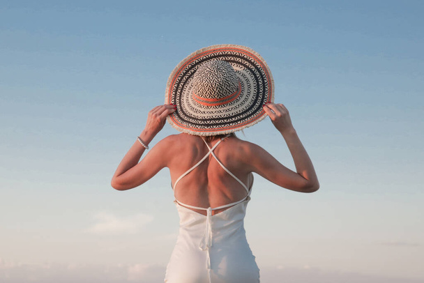 Νεαρή γυναίκα με μακρύ λευκό φόρεμα και ψάθινο καπέλο στην παραλία. Κομψό καλοκαιρινές διακοπές μόδας. - Φωτογραφία, εικόνα