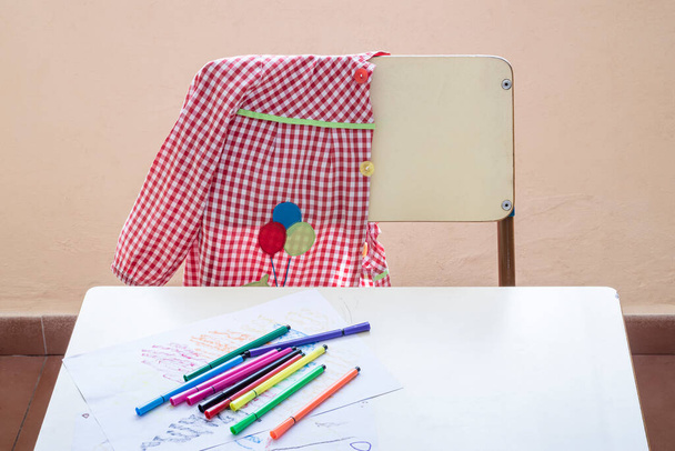 Photo of an elementary school student's place of study.In the photograph you can see a desk, an apron hanging on a chair and some markers on a drawing made by a primary school child. - Photo, Image