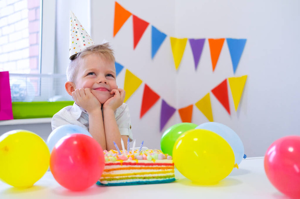 Blonde caucasian boy sits thoughtfully and dreamily at festive table near birthday rainbow cake and makes a wish. Colorful background with balloons - Photo, image