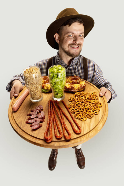 Happy smiling man dressed in traditional Bavarian costume holding round wooden tray with festive food and beer glasses filled with wild hot and barley - Photo, image