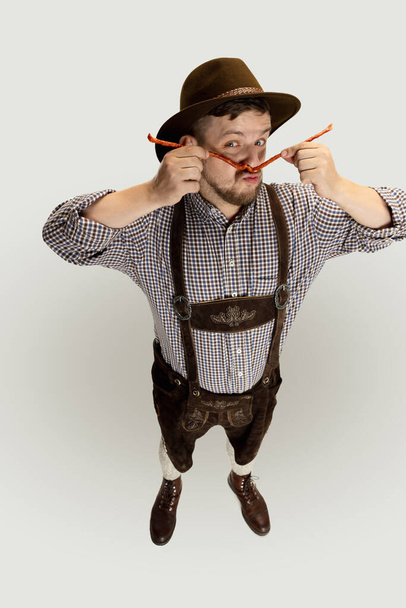 Cute bearded man, waiter in traditional Austrian or Bavarian costume holding smoked sausages isolated over gray background. Funny meme emotions - Fotó, kép