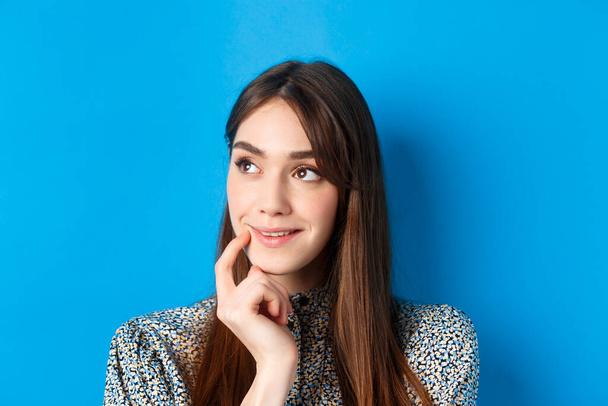 Interesting. Pensive young girl looking aside at logo and smiling, touching lip thoughtful, have an idea, standing on blue background - Фото, зображення