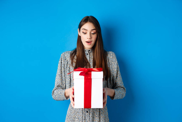 Holidays. Surprised young woman looking at gift with amazed face, receive Valentines day present, standing in dress on blue background - Foto, Imagen