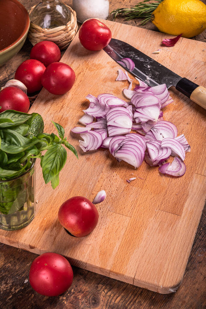 Cocinar ensalada vegetariana. La cebolla en rodajas en la tabla de cortar con los ingredientes para la ensalada vegetal - los tomates, los pepinos, la albahaca, la menta y el limón - Foto, Imagen