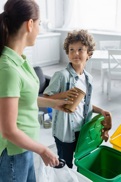 Мальчик берет бумажные стаканчики у матери рядом с мусорным баком с табличкой "Переработка" дома  - Фото, изображение