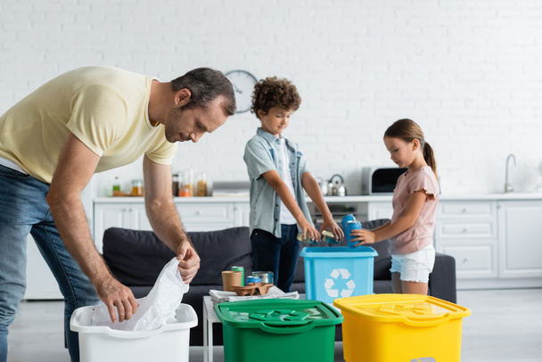 Férfi válogatás szemét közelében elmosódott gyerekek és szemetes dobozok újrahasznosítás tábla otthon  - Fotó, kép