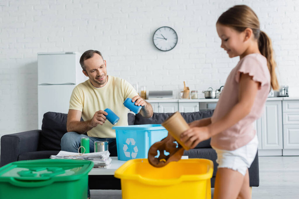 Mosolygó férfi gazdaság ón dobozok közelében doboz újrahasznosítás jel és elmosódott lánya otthon  - Fotó, kép