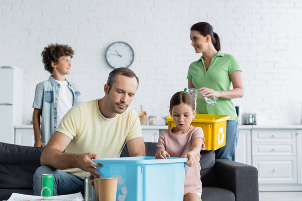 Father and daughter holding box with recycle sign near blurred family in kitchen  - Foto, Bild