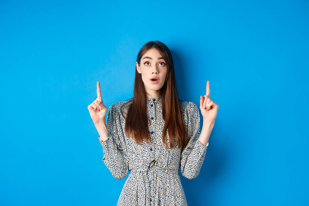 Surprised romantic girl in dress pointing, looking up with opened mouth and fascinated gaze, standing on blue background - Photo, image