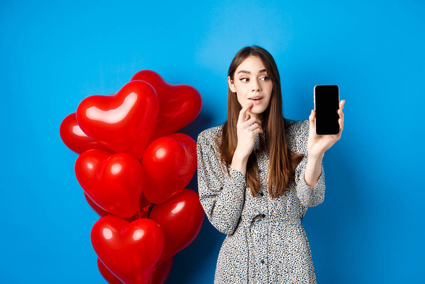 Valentines koncept. Krásná dívka hledá a ukazuje mobilní obrazovku, odhlášení online nákupní nabídka na milence den, stojící na modrém pozadí - Fotografie, Obrázek