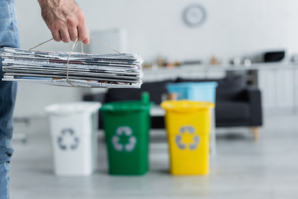 Vista cortada do homem segurando jornais perto de latas de lixo borradas com sinal de reciclagem em casa  - Foto, Imagem