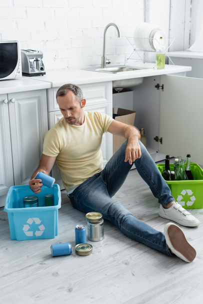 Mężczyzna siedzi w pobliżu pudełek ze znakiem recyklingu i śmieci na podłodze w kuchni  - Zdjęcie, obraz