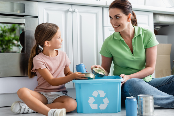 Mosolygó nő és lánya gazdaság ón dobozok közelében doboz újrahasznosítás jel a konyhában  - Fotó, kép