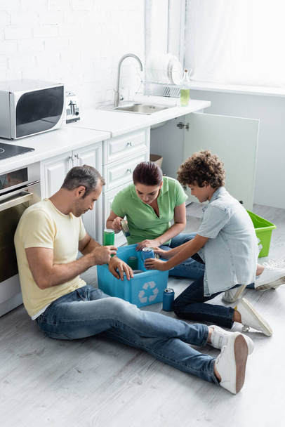 Οικογένεια με γιο βάζοντας τενεκεδάκια σε κουτί με πινακίδα ανακύκλωσης στην κουζίνα  - Φωτογραφία, εικόνα