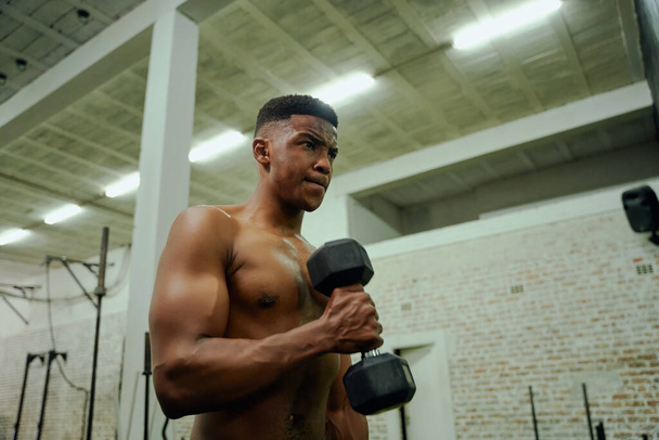 African American male holding weights during an intense workout. Male athlete looking at himself in the gym mirror while lifting dumbbells. High quality photo - Photo, Image