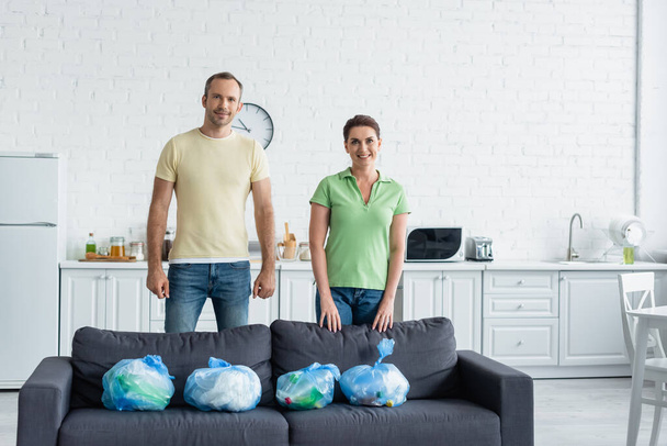 Sorridente coppia guardando la fotocamera vicino sacchetti della spazzatura sul divano in cucina  - Foto, immagini