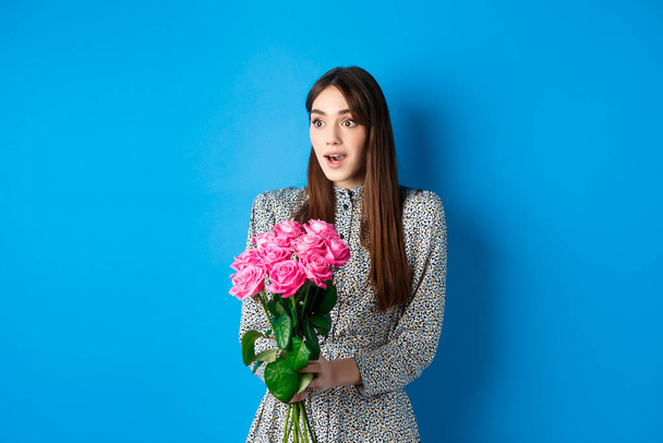 Koncept Valentýna. Překvapená šťastná dívka dívá stranou s úžasem, obdrží kytici romantických květin, drží růžové růže, stojí v šatech na modrém pozadí - Fotografie, Obrázek