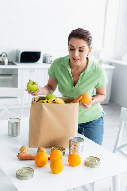 Γυναίκα κρατώντας φρούτα κοντά σε χάρτινη σακούλα στην κουζίνα  - Φωτογραφία, εικόνα