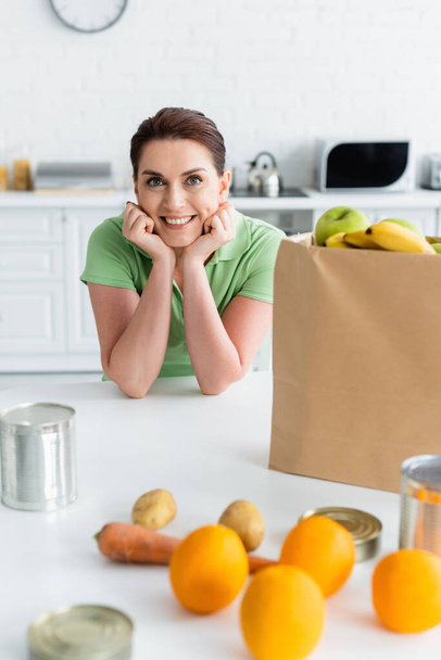 Femme souriante regardant la caméra près de la nourriture floue et sac en papier dans la cuisine  - Photo, image
