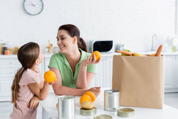 Lächelnde Mutter und Kind halten Orangen in der Nähe von Lebensmitteln und Bastelpapier in der Küche  - Foto, Bild