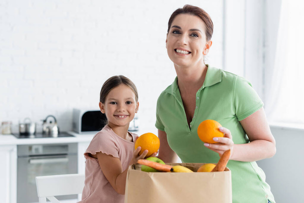 Happy mother and kid standing near paper bag with food in kitchen  - Foto, imagen