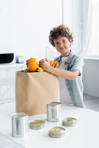 Garçon souriant tenant de la nourriture près du sac en papier et boîtes de conserve dans la cuisine - Photo, image