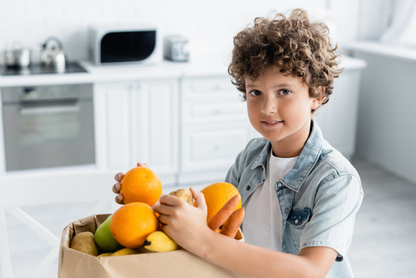 Pozytywne dziecko gospodarstwa owoców w pobliżu torby rzemiosła w kuchni  - Zdjęcie, obraz