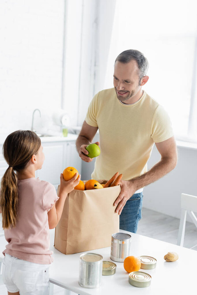 Mosolygós férfi kezében alma közel papírzacskó és lánya a konyhában  - Fotó, kép