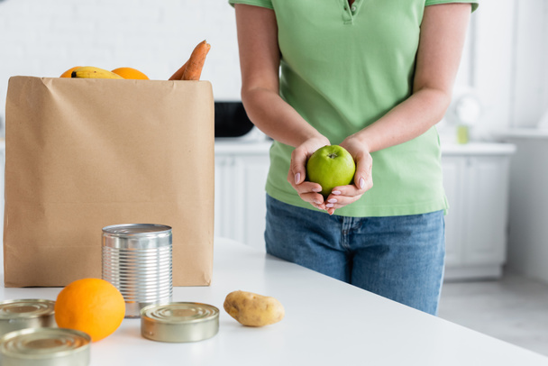 Cropped view of woman holding apple near food and paper bag in kitchen  - 写真・画像