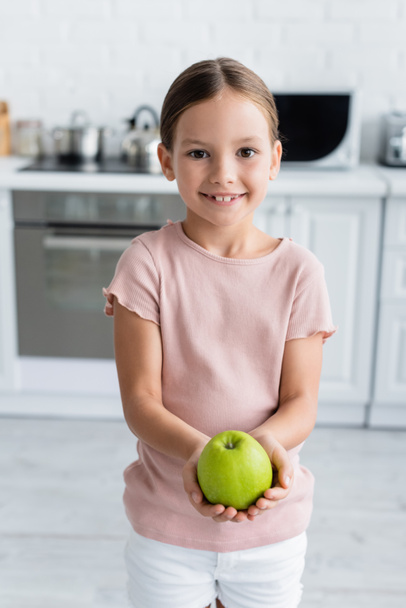 Souriante fille tenant pomme fraîche et regardant la caméra dans la cuisine  - Photo, image