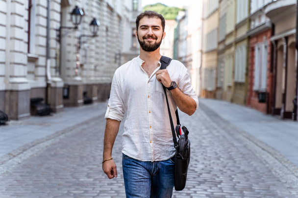 Kráčející pohledný chlap v bílé košili a džíny na pozadí města. Usmívající se muž student nebo kancelářský pracovník - Fotografie, Obrázek