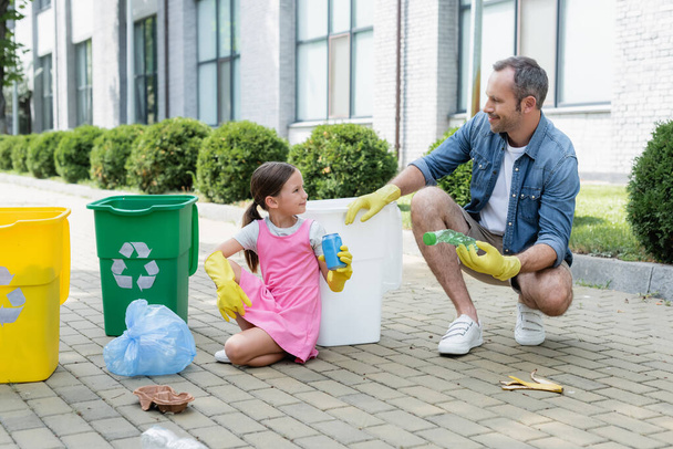 Uśmiechnięta córka i ojciec w gumowych rękawiczkach trzymający śmieci w pobliżu puszek z symbolem recyklingu na zewnątrz  - Zdjęcie, obraz