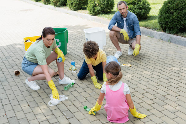Familia con niños en guantes de goma recogiendo basura cerca de latas al aire libre  - Foto, Imagen