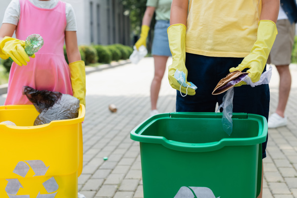 Vista recortada de los niños en guantes de goma clasificando la basura cerca de latas al aire libre  - Foto, imagen