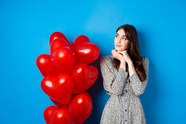 San Valentino. Bella donna sognante in abito, imaging appuntamento romantico, in piedi vicino a palloncini cuori rossi e sorridente, in piedi su sfondo blu - Foto, immagini