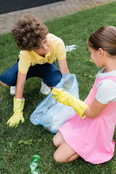 Kids in rubber gloves putting trash in bag on grass  - Фото, изображение