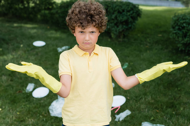 Verwirrter Junge in Gummihandschuhen blickt in Kamera in der Nähe von verschwommenem Müll im Freien  - Foto, Bild