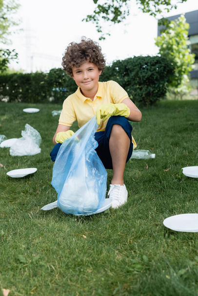 Niño sonriendo a la cámara mientras sostiene la bolsa de basura cerca de la basura en el césped  - Foto, Imagen
