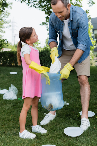 Smiling girl holding bag near father with trash outdoors  - Fotoğraf, Görsel