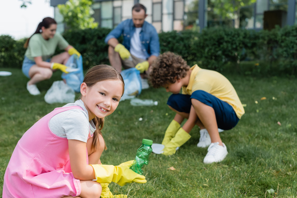 Smiling girl holding bottle near blurred brother and parents collecting trash outdoors  - Photo, Image