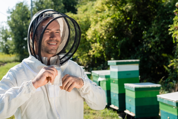 ευτυχισμένη μελισσοκόμος προσαρμογή προστατευτική στολή στο μελισσοκομείο - Φωτογραφία, εικόνα