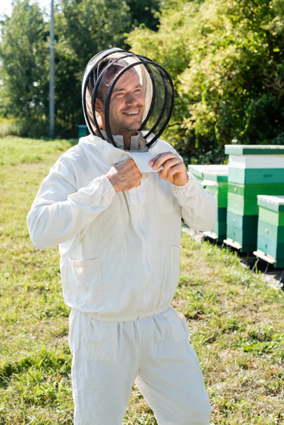 smiling apiarist adjusting beekeeping suit near beehives on apiary - Photo, Image