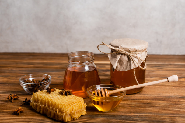 jars with honey near honeycomb, anise seeds and dipper on wooden table and grey background - Photo, Image