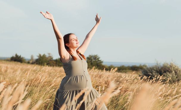 young beautiful pregnant woman walks in field among dry fluffy grass enjoying sunset, relaxation on summer nature, preparation for childbirth and happy, healthy motherhood - Foto, immagini