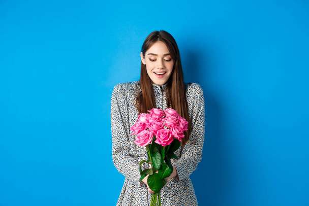 Concetto di San Valentino. Felice donna attraente ricevere fiori a sorpresa, guardando grato al bouquet di rose rosa, in piedi su sfondo blu - Foto, immagini