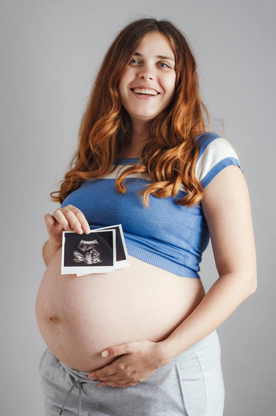 smiling pregnant cute young caucasian woman with red hair and blue eyes standing against grey studio background and holding an ultrasound black and white scan, happy mother expectant - Foto, Imagem