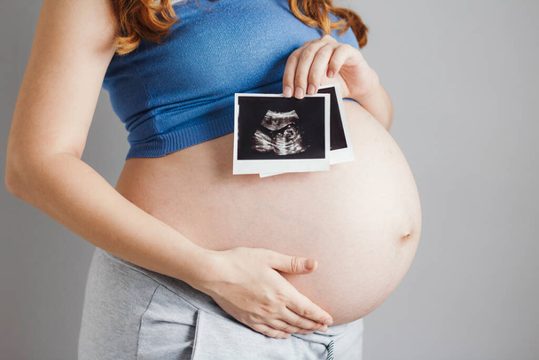 smiling pregnant cute young caucasian woman with red hair and blue eyes standing against grey studio background and holding an ultrasound black and white scan, happy mother expectant - Photo, image