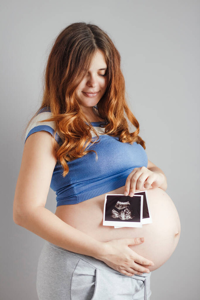 smiling pregnant young woman with red hair and blue eyes standing against grey studio background and holding an ultrasound black and white scan, happy mother expectant - Foto, Bild