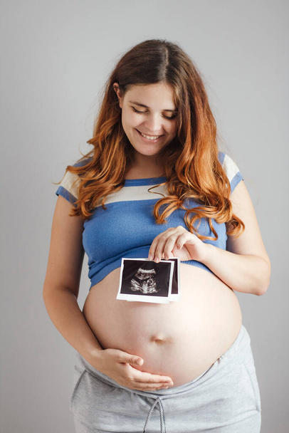 smiling pregnant young woman with red hair and blue eyes standing against grey studio background and holding an ultrasound black and white scan, happy mother expectant - Photo, Image