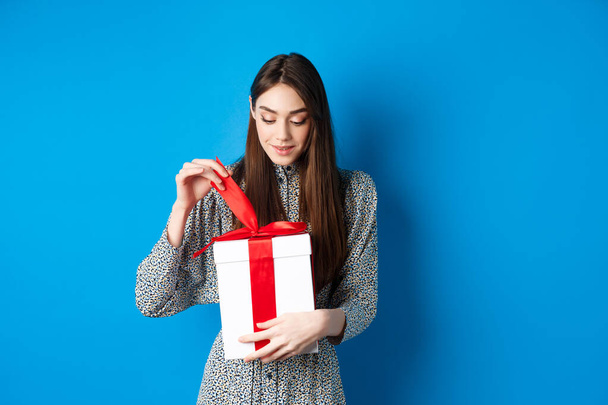 День Святого Валентина. Милая молодая женщина открывает коробку с подарком, снять ленту из настоящего и улыбаясь заинтригованы, стоя на синем фоне - Фото, изображение
